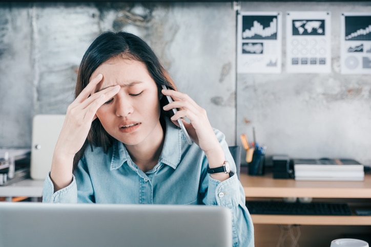 Stressed Asian creative designer woman cover her face with hand and feel upset while talk on mobile phone with customer in front of laptop computer on desk at office,Stress office lifestyle concept.