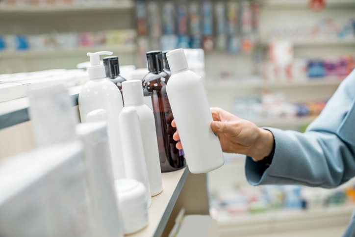 Woman taking a bottle with cosmetics from the shelf of the pharmacy supermarket, close-up view on the bottle with blank label