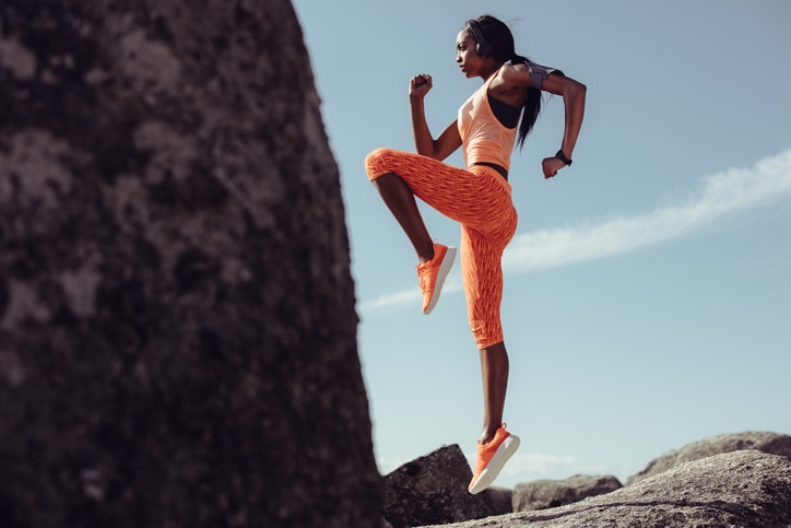 Sports woman doing jumping and stretching workout outdoors. Full length of healthy young african fitness woman doing exercise at rocky beach.