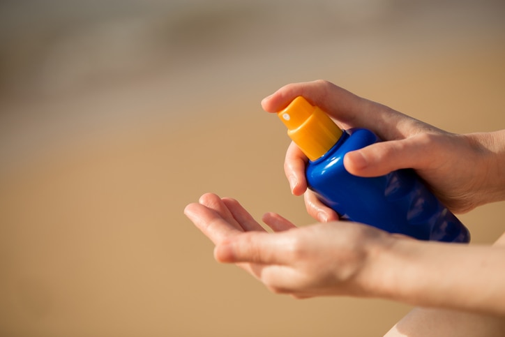 Young woman sitting on sand seashore, holding bottle of sunscreen lotion before applying, close up of hands