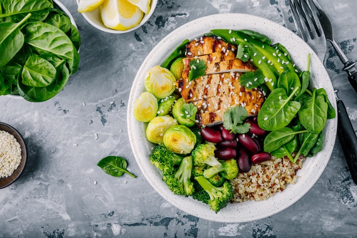 Healthy buddha bowl lunch with grilled chicken, quinoa, spinach, avocado, brussels sprouts, broccoli, red beans with sesame seeds on dark gray background. Top view.