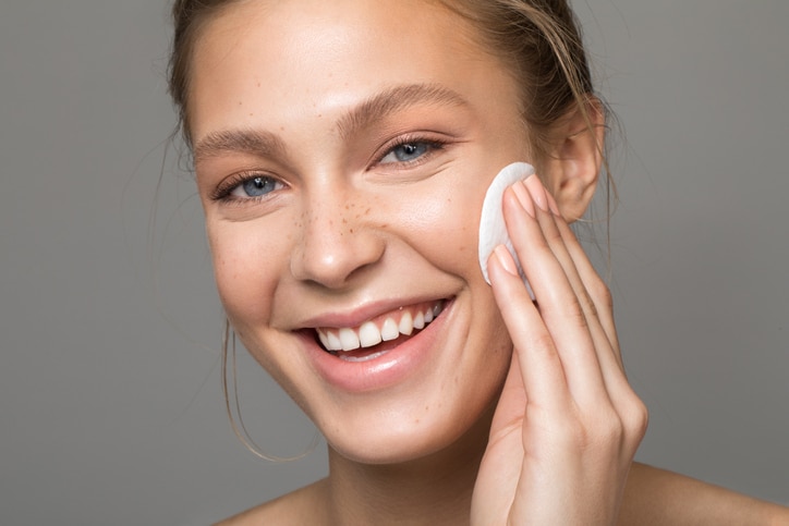 Closeup studio shot of a beautiful young woman with freckles skin, cleansing her face with cotton pad. Posing against a grey background