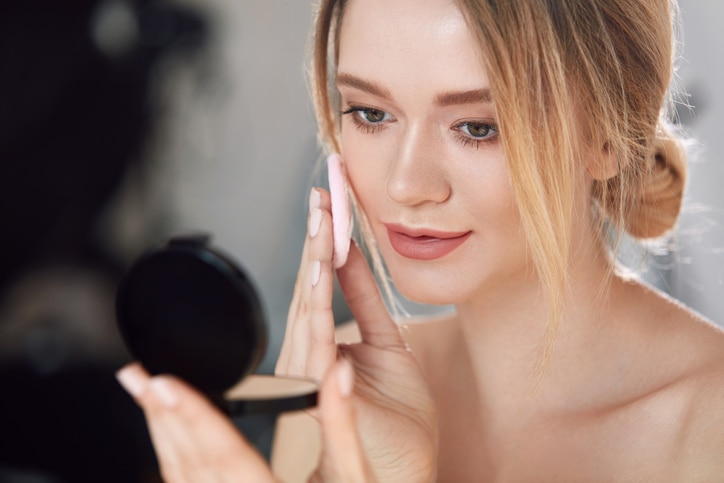 Face Make-Up. Closeup Of Sexy Female Applying Dry Powder Foundation Looking In Mirror. Portrait Of Young Woman Putting Makeup Powder With Cosmetic Cushion On Her Facial Skin Indoors. High Resolution