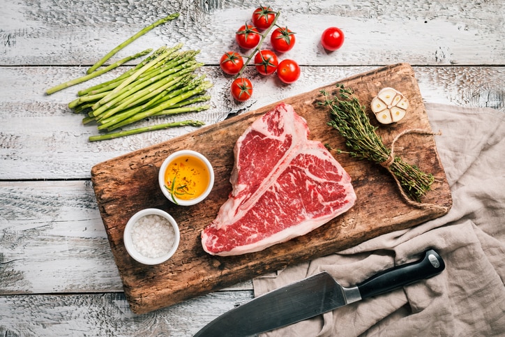 Raw T-bone Steak for grill or BBQ on aged cutting board over white wooden background, top view