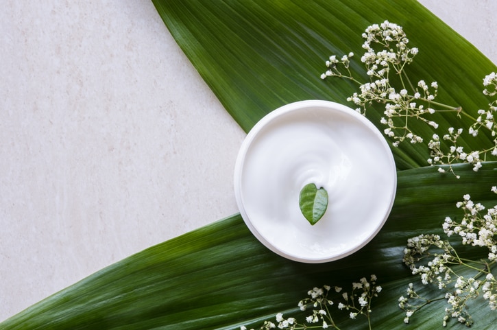 Top view of cosmetic lotion with white flowers and green leaf. Skin care beauty treatment with jar of body moisturizer. High angle view of white body lotion with little green leaf on marble background.