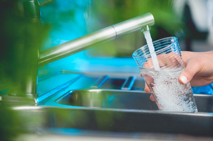 Woman filling a glass of water. She is using the faucet in the kitchen sink. There is a plant out of focus in the foreground. Close up with copy space.