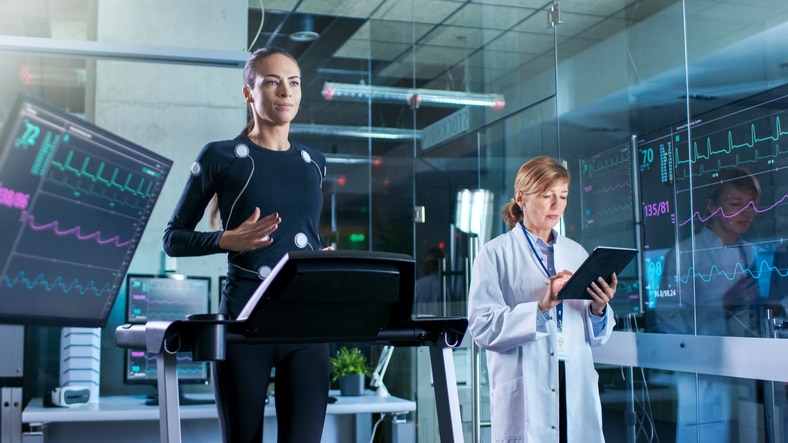 Woman Athlete Walks on a Treadmill  with Electrodes Attached to Her Body while Scientist Holding Tablet Computer Supervises whole Process. In the Background Laboratory with Monitors Showing EKG Readings.