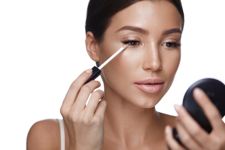 Beauty Face. Closeup Of Sexy Young Woman With Concealer Brush And Mirror In Hands. Portrait Of Beautiful Healthy Female Model Applying Concealer Under Eyes. Skin Cosmetics. High Resolution