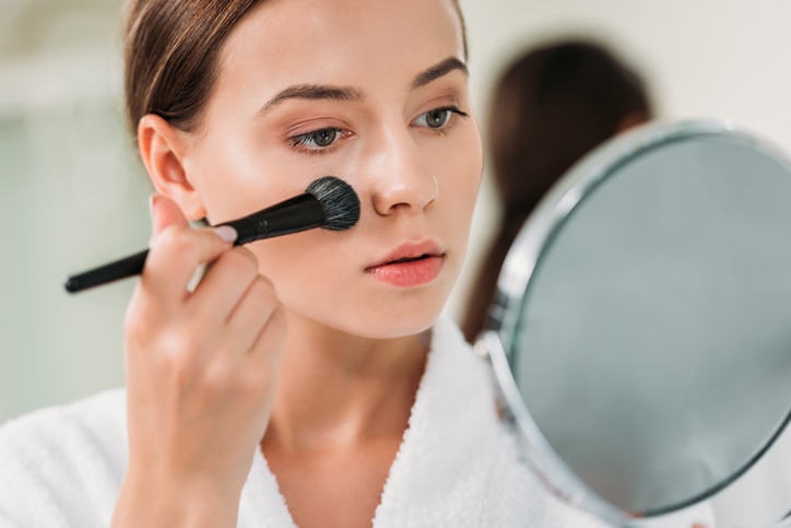 beautiful young woman in bathrobe applying makeup and looking at mirror