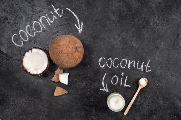 Top view of coconut oil in glass jar with spoon and cracked coconut on black