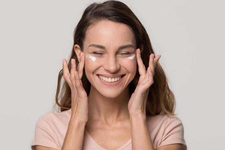 Smiling happy pretty young teen girl woman applying moisturizing lotion cosmetic cream on face for clean healthy soft skin care natural beauty treatment isolated on white grey studio blank background