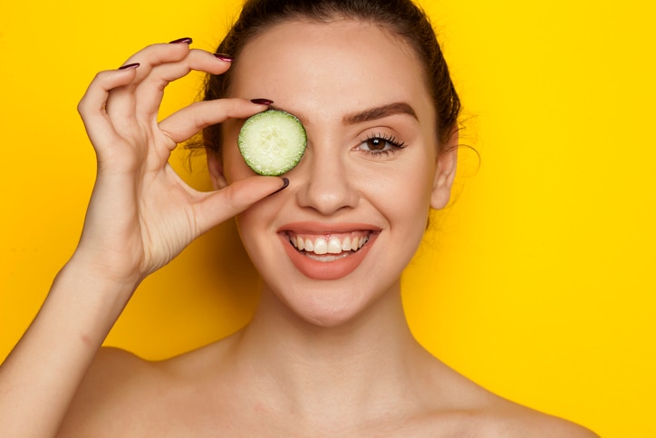 Happy young woman posing with slices of cucumber on her face on yellow background