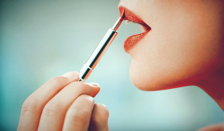 Closeup side view of unrecognizable woman putting on red lipstick with lip brush. Shot against cyan green blurry background.