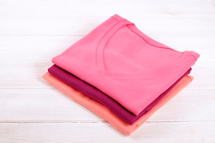 Stack of colorful perfectly folded v neck t-shirts on white wooden texture table background. Pile of different pastel color shirts and sweaters. Background, close up, top view, copy space for text.