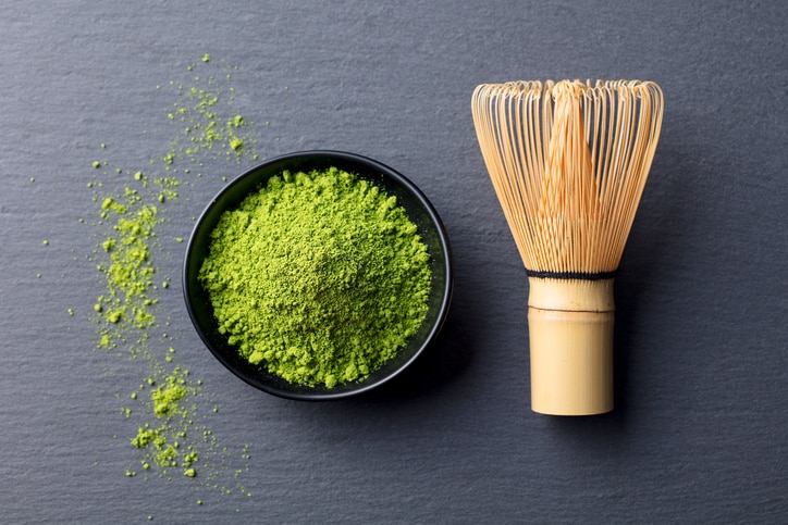 Matcha, green tea powder in black bowl with bamboo whisk on slate background. Top view