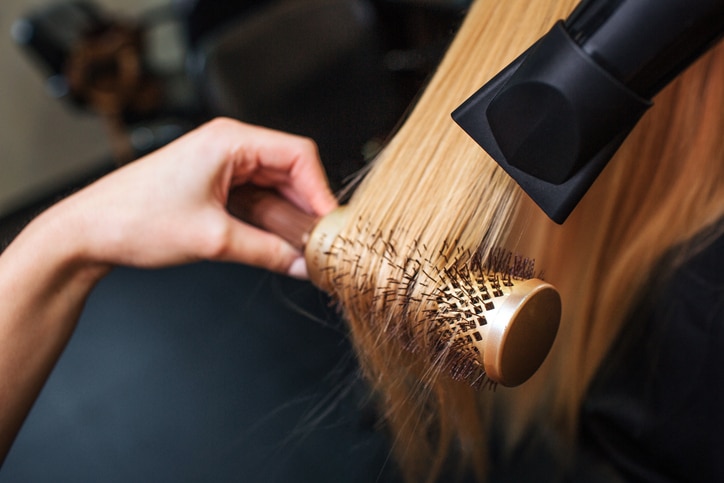 Female client in beauty salon. Close-up of hairdressers hand drying blond hair with hair dryer and round brush, doing professional hairstyle