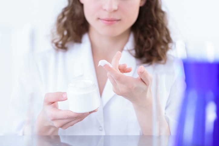 Female chemist in laboratory coat holding a white container and presenting lotion on her finger