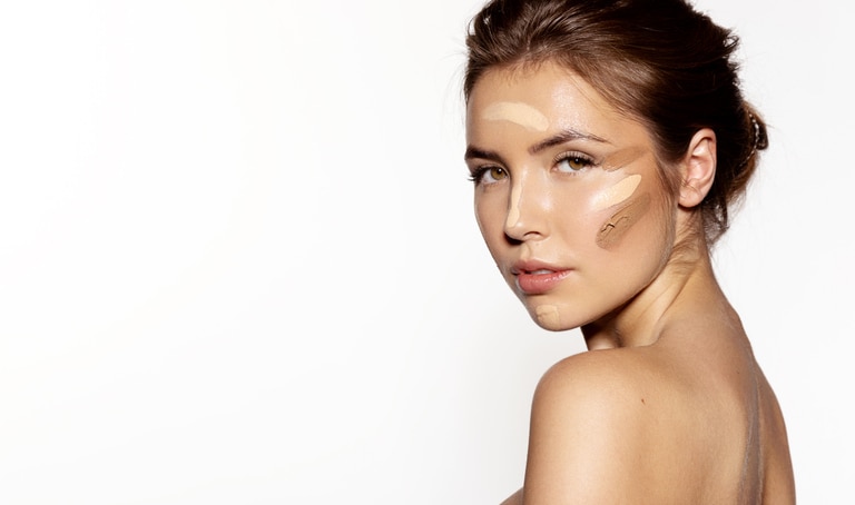 Waist up portrait of sensual young woman with different kinds of foundation on her face. Cute girl with maquillage posing at studio. Beauty treatment concept. Isolated on grey background