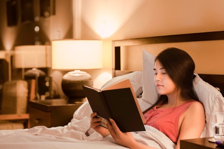 Beautiful biracial Asian Caucasian teen girl reading book  in bed at night with yellow lamp light on walls