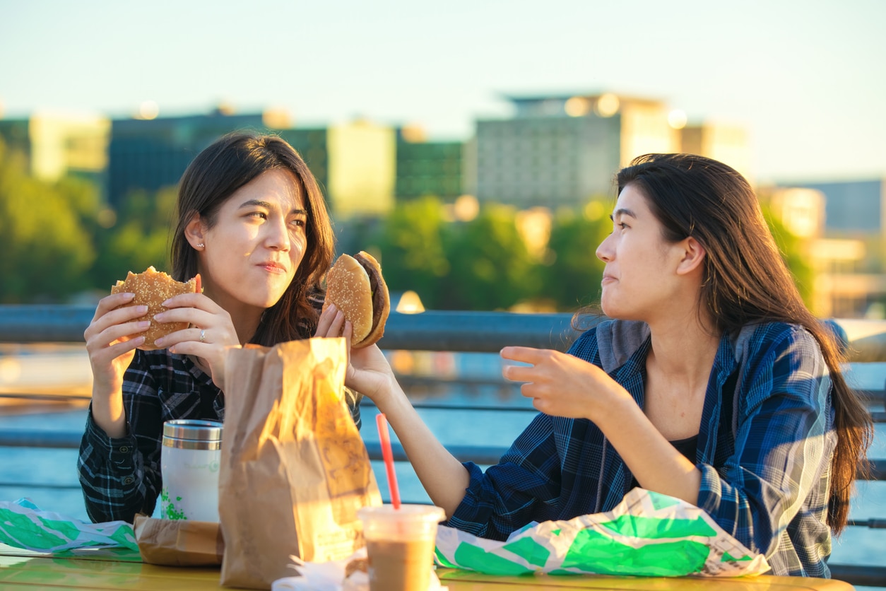 Two biracial Asian, Caucasian  teen girls  or youmg women eating hamburgers outdoors at sunset by lake in urban area, talking and smiling