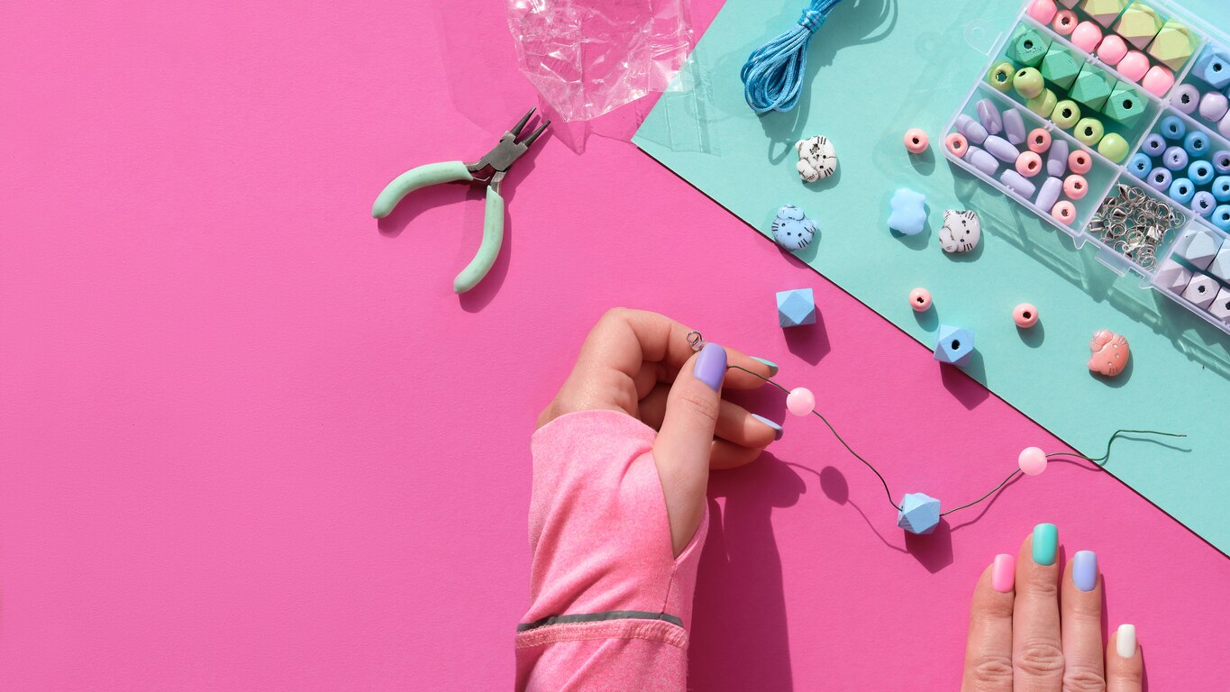 Handmade jewelry. Box with beads, hands with tools. Making handmade jewelry for yourself or as self made custom gifts. Flat lay, top view on split pink and mint green paper background.