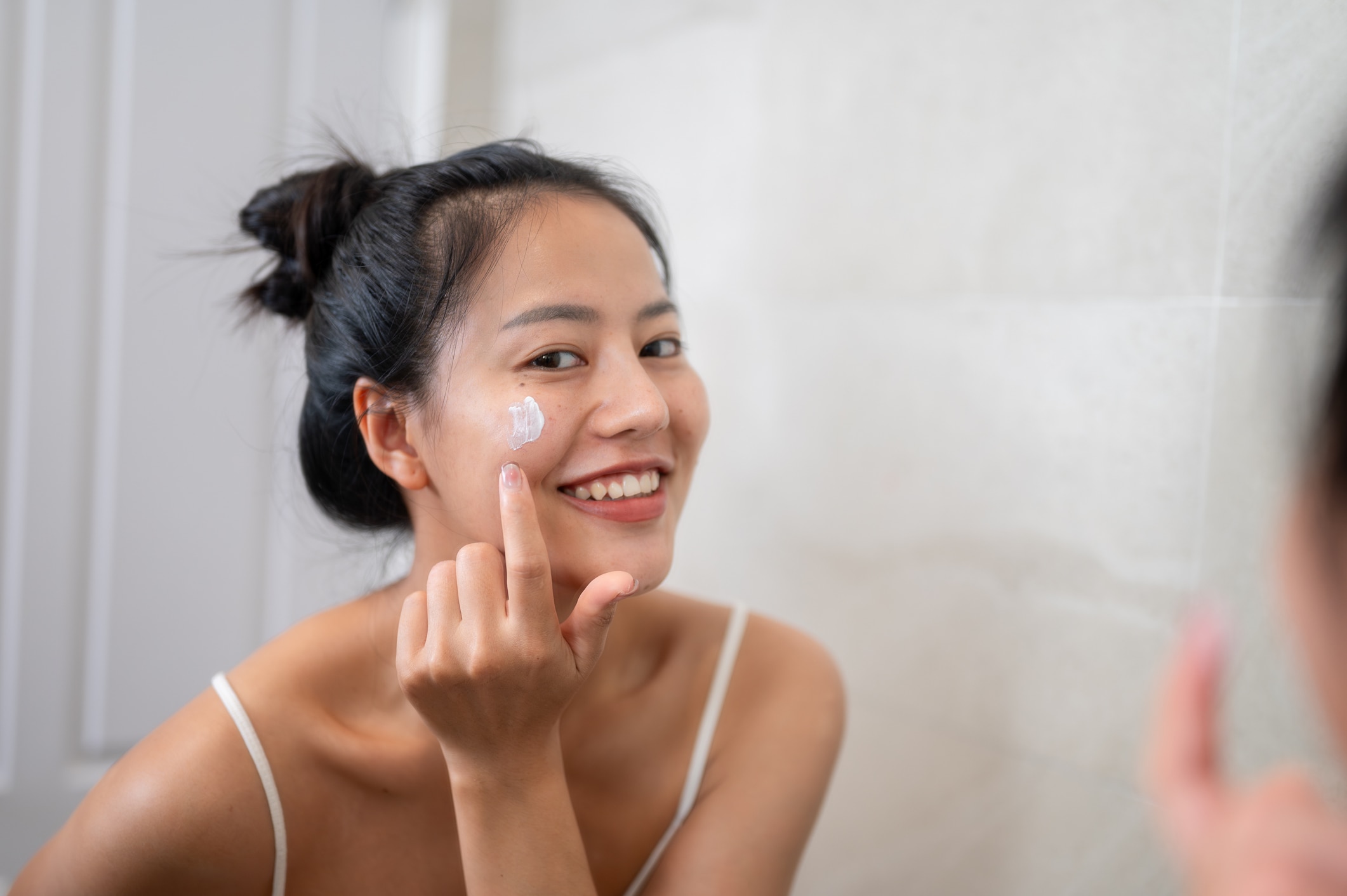 An attractive Asian woman is smiling at the mirror while applying facial cream to her face.
